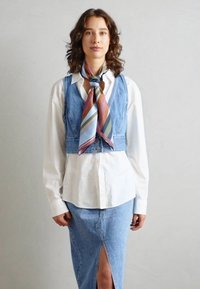 Gilet in denim sopra una maglietta bianca, abbinato a una gonna in denim con uno spacco frontale. Una sciarpa in seta multicolore aggiunge dettagli.