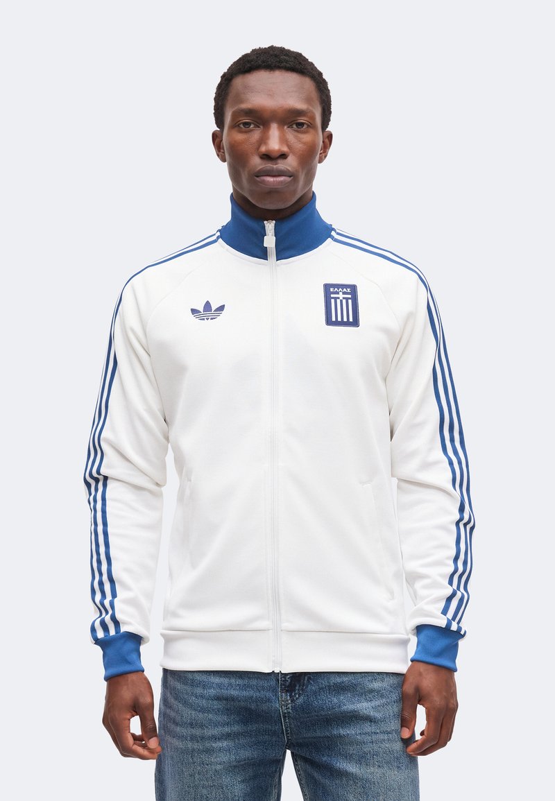 Άνδρας που φοράει λευκό αθλητικό μπουφάν Adidas με μπλε ρίγες και ψηλό γιακά, συνδυασμένο με μπλε τζιν, στέκεται μπροστά σε φωτεινό φόντο.