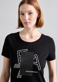 Čierne bavlnené tričko s bielym grafickým dizajnom, vrátane textu "EA" a obdĺžnikového čierneho akcentu s logom "Emporio Armani".