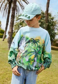 Felpa azzurra chiara con stampa tropicale vivace, caratterizzata da palme e figure giocose. Abbinata a un cappellino azzurro chiaro e jeans strappati.