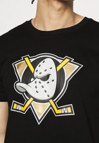 Person som bär en svart tröja med en stiliserad maskerad anka ansikte och korsade gula hockeyklubbor bakom.