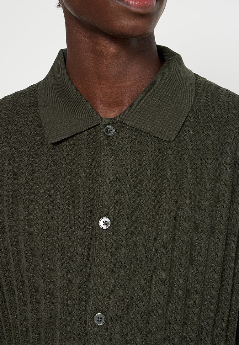 Dunkelgrünes Strickhemd mit gerippter Struktur, ausgestattet mit einem umschlagbaren Kragen und schwarzen Knöpfen. Nahaufnahme des oberen Teils, die Designdetails hervorhebt.