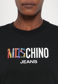 Juoda marškinėliai su grafika, kurioje spalvoti užtrauktukai sudaro tekstą "KIDS CHINO JEANS" baltais rašmenimis. Sklandi audinio tekstūra.