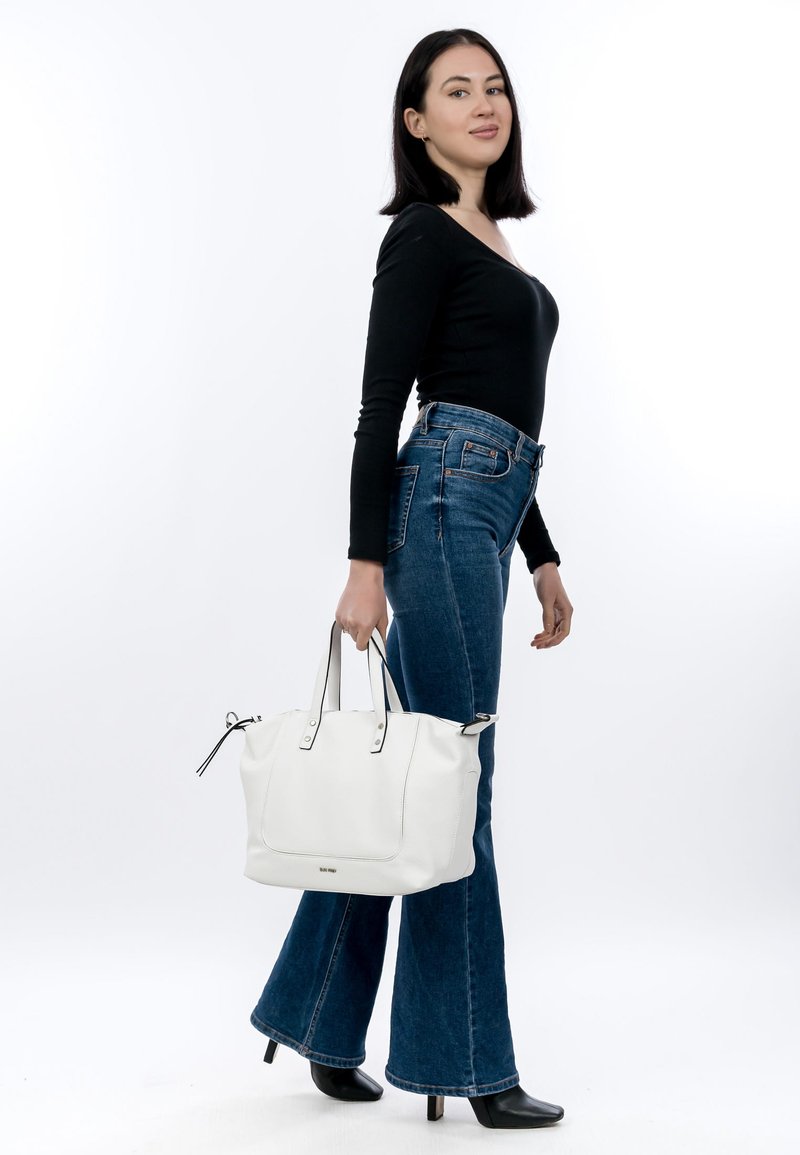 Kvinde iført sort top med lange ærmer og vide jeans, der holder en hvid lædertaske, stående i et studie med en ensfarvet hvid baggrund.