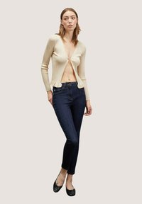 Cárdigan beige de canalé con un profundo escote en V y dobladillo con volantes, llevado sobre unos jeans skinny azul oscuro. La modelo está de pie con zapatos planos negros sobre un fondo neutro.