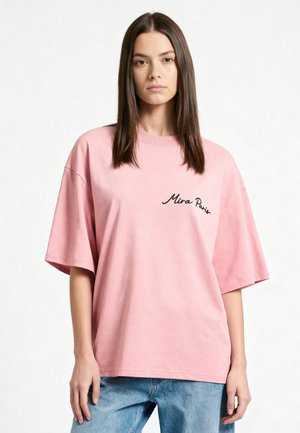 Jeune femme aux cheveux bruns raides, portant un t-shirt rose ample avec le texte « Mira Paris » et un jean bleu, debout devant un fond uni.