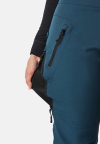 Blaue technische Hose mit einer Reißverschlusstasche, Mesh-Futter und schwarzen Akzenten. Das Material wirkt leicht und robust. Der Zipper-Anhänger ist sichtbar.
