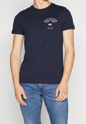 Homme portant un t-shirt Hilfiger New York bleu marine avec logo et un jean bleu clair, debout les bras détendus le long du corps.