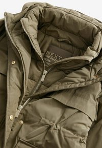 Jachetă puffer de culoare verde măslinie cu un design matlasat, fermoar frontal și căptușeală în două tonuri. Are o etichetă în interior cu numele brandului și logo-ul.