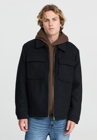 Giacca nera in misto lana con due tasche frontali, chiusura con zip e vestibilità leggermente over size, indossata sopra un hoodie marrone e jeans blu.