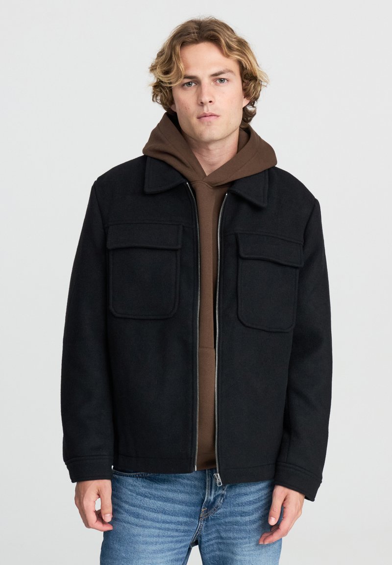 Giacca nera in misto lana con due tasche frontali, chiusura con zip e vestibilità leggermente over size, indossata sopra un hoodie marrone e jeans blu.