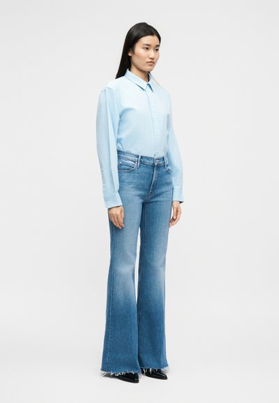 Mladá žena oblečená v svetlomodrej košeli na gombíky a vysokých rozšírených modrých džínsoch s opranými lemami, stojaca na jednoduchom pozadí.