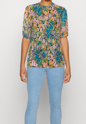 Femme portant un chemisier à manches courtes à fleurs multicolores associé à un jean bleu clair, debout devant un fond blanc uni.