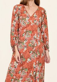Lång, rostfärgad klänning med blommönster i vitt, gult och grönt. V-ringning, långa puffärmar, texturerat tyg och sidofickor.