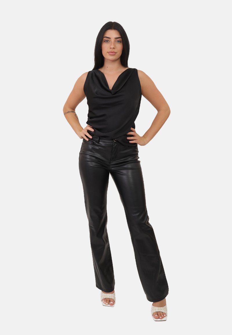 Una donna con lunghi capelli scuri sta in piedi con sicurezza indossando un top drappeggiato senza maniche nero, pantaloni in pelle neri e sandali con tacco bianchi su uno sfondo bianco.