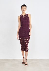 Robe tricotée bourgogne avec découpes ovales, sans manches, longueur mi-mollet et silhouette ajustée, associée à des talons noirs à lanières.