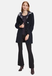 Navy toggle coat with a plaid-lined hood, featuring a tailored fit, wool blend fabric, and black trousers paired with heeled boots.
