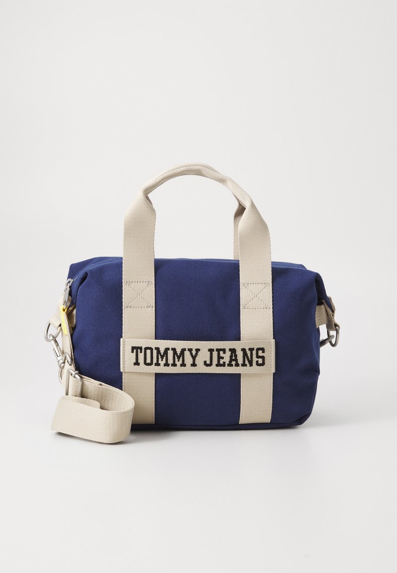 Modrá látková taška s krémovými uchy a popruhem, s výrazným logem "TOMMY JEANS". Kompaktní obdélníkový tvar s zipem a kovovými detaily.