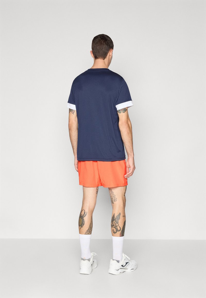 Marine t-shirt med hvide accenter og orange shorts. Atletisk pasform, letvægts stof, synlige tatoveringer på benene, hvide sokker og sneakers.