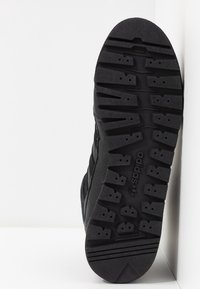 Semelle de basket noire en caoutchouc texturé, avec des rainures profondes et des trous pour l'adhérence. Marquage visible au-dessous, bords lisses.