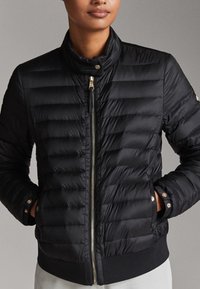 Veste en doudoune noire avec un design matelassé, col montant et accessoires en or. Équipée de poignets et d'une taille côtelés pour un look ajusté.