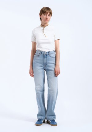 Camiseta blanca de manga corta, jeans de tiro alto de color azul claro y zapatillas azules. La modelo lleva un collar tipo choker y tiene el cabello corto y castaño.