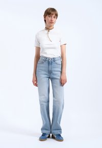 Bílý top s krátkým rukávem, světle modré džíny se zvýšeným pasem a modré tenisky. Model má na sobě choker náhrdelník a krátké hnědé vlasy.