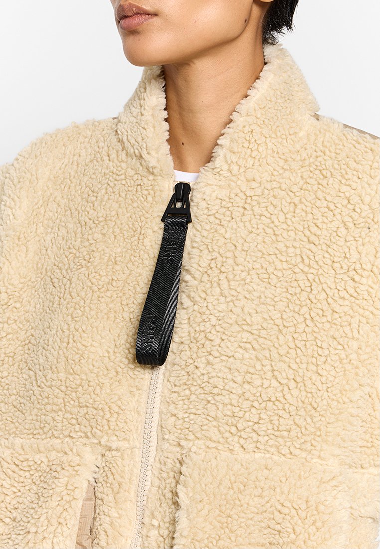 Persona che indossa una giacca di pile beige, testurizzata, con un collo alto e un'impugnatura nera della zip visibile.