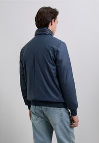 Marinblå syntetisk jacka med hög krage, ribbade muddarna och en slät yta. Har en minimalistisk design, utan synliga mönster eller hårdvara.