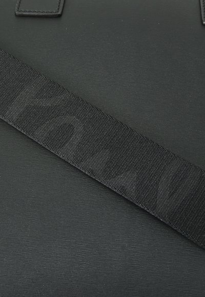 Black bag made of textured material with a wide black strap featuring a subtle logo pattern. Includes a square leather patch.
