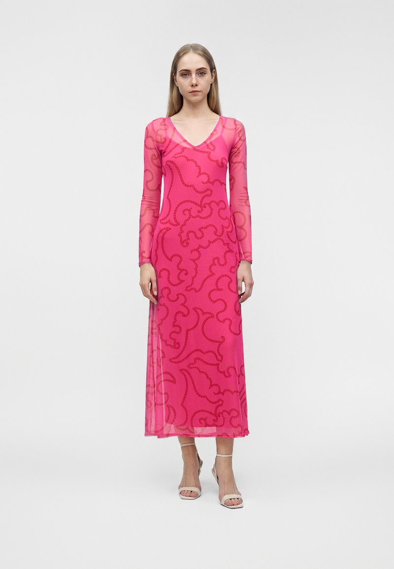 Jeune femme debout, portant une longue robe rose avec des motifs rouges abstraits, manches longues transparentes et talons ouverts blancs.