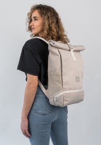 Fiatal nő, göndör hajjal, fekete pólót és kék farmernadrágot visel, nagy bézs színű, oldalsó zsebekkel ellátott roll-top hátizsákot hord.