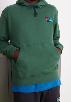 Sudadera verde con un bolsillo frontal y capucha ajustable, que presenta un parche de logo rojo y azul. Lleva puesta sobre una camiseta azul y pantalones negros.