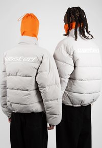Chaquetas de plumas grises con diseño acolchado y puños elásticos. Logotipo "Wasted" en blanco en la parte posterior. Modelo con un cubierta naranja en la cabeza.