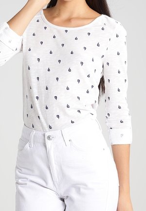 Top bianco a maniche lunghe con una piccola stampa di velieri blu navy, scollatura rotonda e maniche arrotolate, abbinato a pantaloni bianchi. Tessuto morbido e leggero.