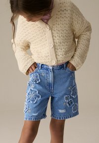 Cardigan crème tricoté, boutonné, sur un haut rose. Shorts en denim bleu avec broderie florale et détails effilochés. Style décontracté, coupe ample.