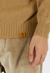 Maglione a coste beige con finitura testurizzata, dotato di un'etichetta quadrata con il logo Timberland in basso a sinistra.