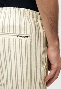 Μπεζ παντελόνι με λεπτές κάθετες μαύρες ρίγες, ελαστική μέση, πίσω τσέπη με ετικέτα "JOOP! JEANS" και ορατό πλευρικό φερμουάρ δίπλα σε χαλαρό χέρι.