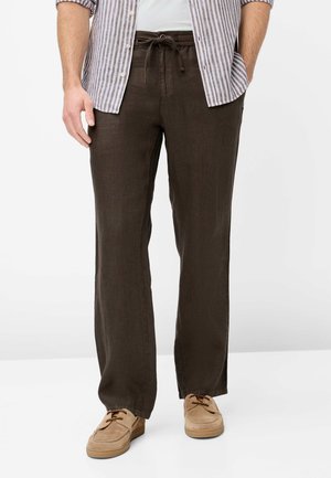 Mann trägt braune Kordelzughose, beige Mokassins und ein gestreiftes Hemd mit Knöpfen und hochgekrempelten Ärmeln.