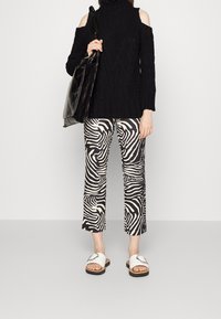 Personne portant un pull noir à épaules dénudées, pantalon imprimé zèbre noir et blanc, sandales blanches à enfiler, tenant un sac cabas en cuir noir.