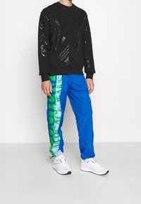 Černá mikina se zembossovaným textem, spárovaná s modrými kalhotami s zeleným grafickým potiskem. Bílé tenisky dokončují celý outfit.