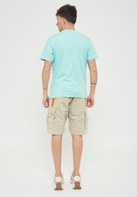 T-shirt di cotone turchese con collo rotondo, abbinata a pantaloni cargo beige con tasche laterali e strisce rosse-gialle. Sneakers con suola color sabbia.