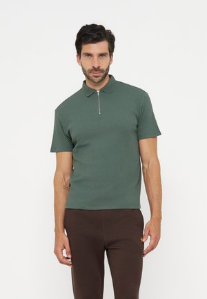 Homme aux cheveux foncés et à la barbe, portant un polo vert à manches courtes avec fermeture éclair et un pantalon marron foncé, debout sur un fond blanc.