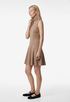 Lyshåret kvinde iført ærmeløs beige rullekraveskjole med udvidet nederdel og sorte Mary Jane-flade sko, stående sideværts mod hvid baggrund.