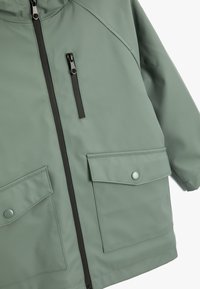 Vattentät grön jacka med en slät textur, med dragkedja fram, två stora fickor med lock och hög krage.