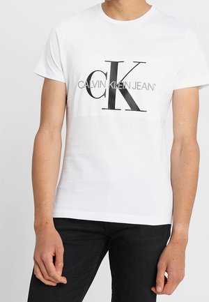 Homme portant un t-shirt blanc Calvin Klein Jeans avec un logo noir et gris à l'avant et un pantalon noir, sur un fond uni.