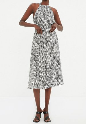 Vrouw die een mouwloze midi-jurk met zwart-wit patroon en halterneck draagt, met zwarte hoge hakken, staande met haar handen in haar taille.