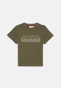 Alyvuogių žalios spalvos medvilnės marškinėliai trumpomis rankovėmis, su dideliu, matinio paviršiaus "HUGO" logotipu centre, esantį ant tekstūruoto stačiakampio fono.