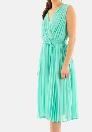 Femme portant une robe plissée sans manches, longueur genou, de couleur vert menthe, avec un col en V et une ceinture nouée à la taille, sur un fond uni.