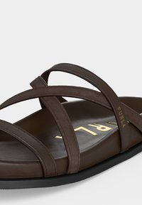 Καφέ δερμάτινο σανδάλι με επίπεδη σόλα, λουριά που διασταυρώνονται και χρυσό λογότυπο "FURLA" στο πέλμα και στο λουράκι, μαύρη σόλα, μινιμαλιστικός σχεδιασμός.